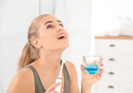 Woman in bathroom holding electric toothbrush and blue mouthwash, preparing to rinse