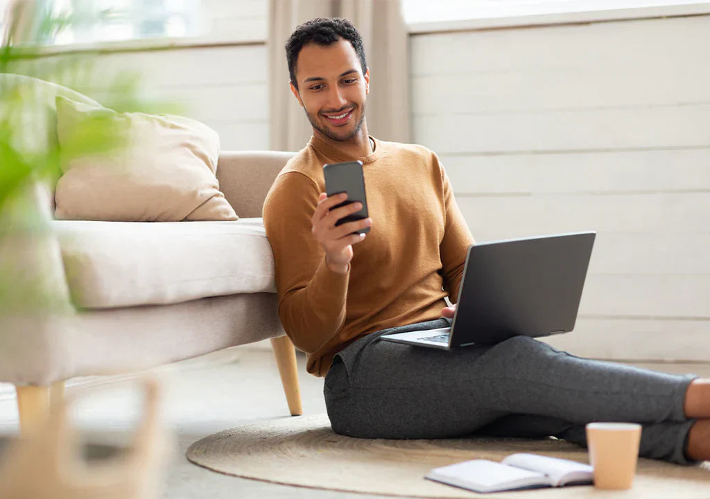 Man working from home on laptop, smiling at smartphone in cozy living room setting