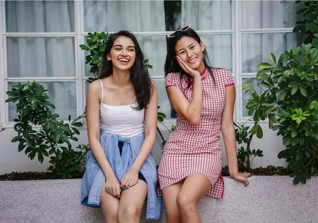 Two smiling women sit outdoors with greenery and window backdrop, casual and summer fashion