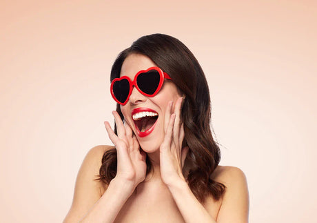 Smiling woman with red heart-shaped sunglasses and red lipstick on beige background